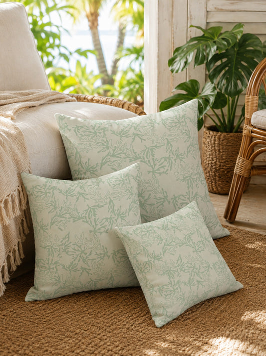 A set of 3 square throw pillows featuring a light sage green coral branch pattern on an off-white background, resting on the ground next to a couch in a tropical living room.