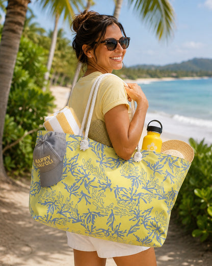 A woman on a tropical beach path carrying a large yellow and sea coral print tote bag with white rope handles. The bag has a gray cap on the side with 'HAPPY' printed in gold, and she is also carrying a yellow water bottle and a straw hat.