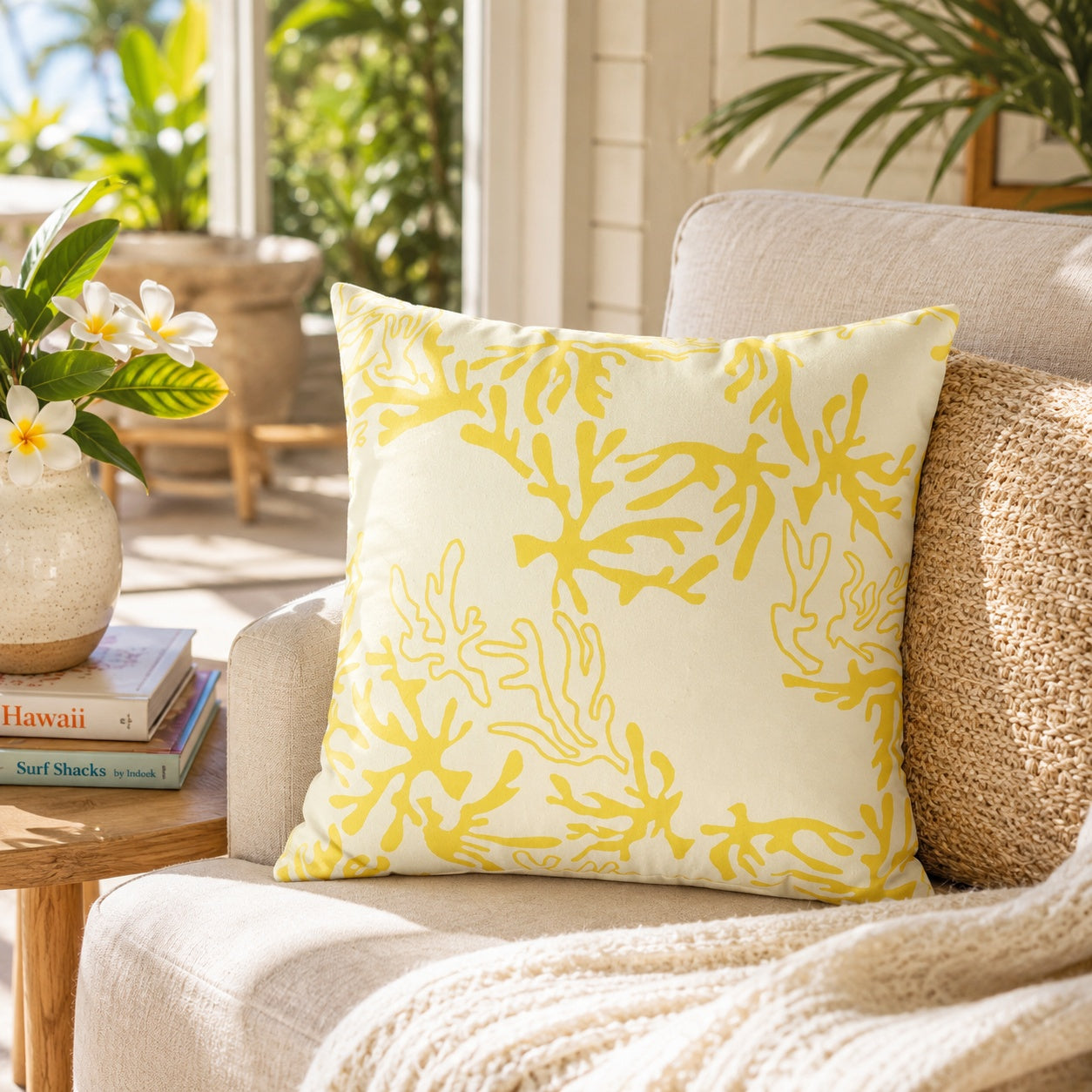 A square throw pillow with a yellow coral branch pattern on a cream background, styled on a beige armchair next to a woven pillow in a bright, tropical-themed room.