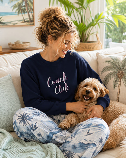 A woman sitting on a white couch with a small fluffy dog, wearing a navy blue sweatshirt that says 'Couch Club' in white script and blue-and-white palm tree print pajama pants.
