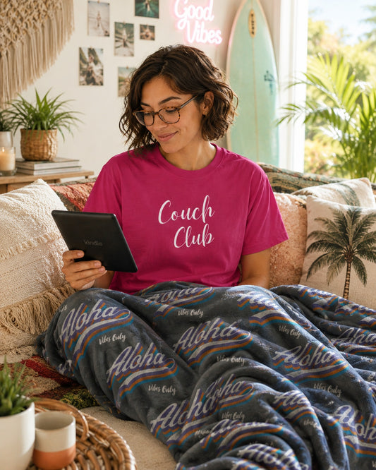 A woman wearing glasses and a pink t-shirt with 'Couch Club' printed on it sits on a couch in a cozy living room, reading a tablet while covered by a gray aloha patterned blanket.