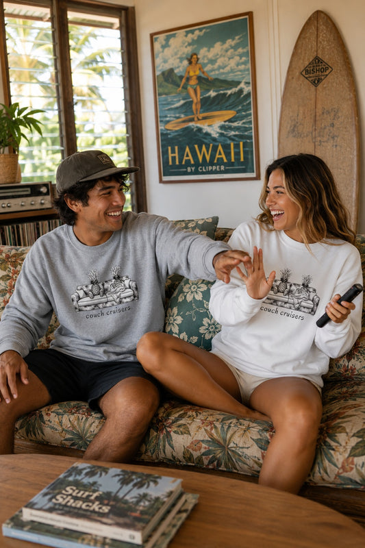 A man and a woman sitting on a floral couch in a living room, both wearing matching graphic crewneck sweatshirts with pineapples sitting on a couch and text 'couch cruisers'. The man wears a gray sweatshirt and a cap, while the woman wears a white sweatshirt.