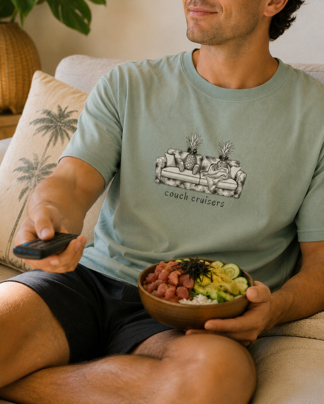 A man sitting on a beige couch holding a bowl of poke food and a remote control, wearing a sage green graphic t-shirt with a pineapple couch design and the text 'couch cruisers'.