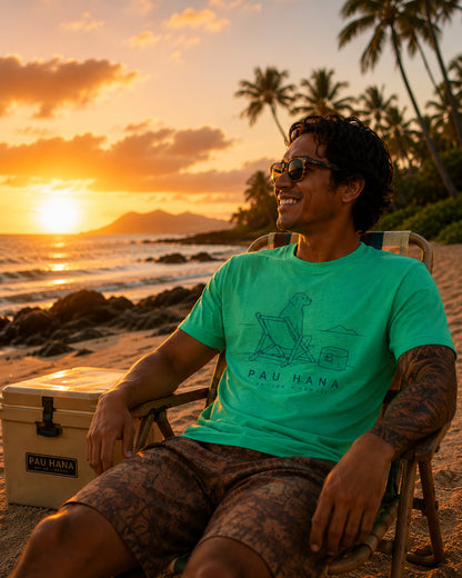 Man sitting on a beach chair watching the sunset wearing a Green t-shirt with a graphic design and 'PAU HANA' text