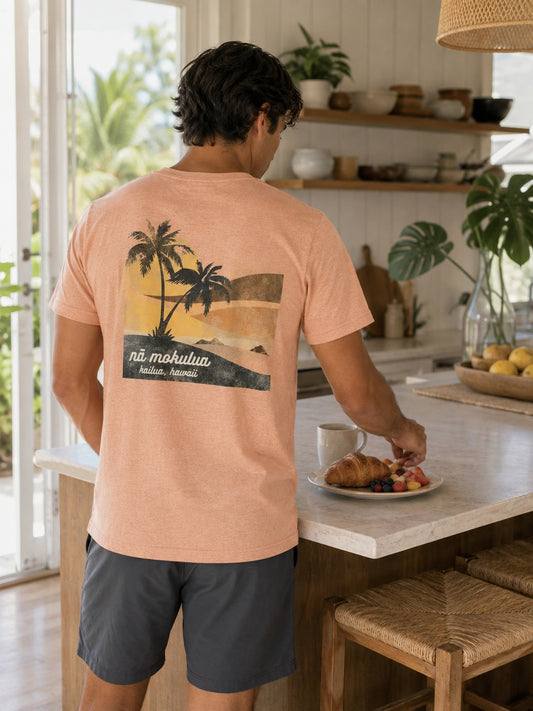 A man standing in a bright kitchen, viewed from behind, wearing a peach-colored t-shirt with a graphic print of palm trees and the text 'na mokulua kailua, hawaii' on the back. He is wearing dark gray shorts and reaching for a croissant on a plate on the kitchen island.