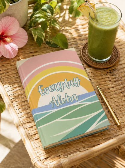 A hardcover notebook with a colorful retro sunset design and the words 'Everyday Aloha' on the cover, styled on a woven rattan tray next to a green drink, a gold pen, and a pink hibiscus flower.