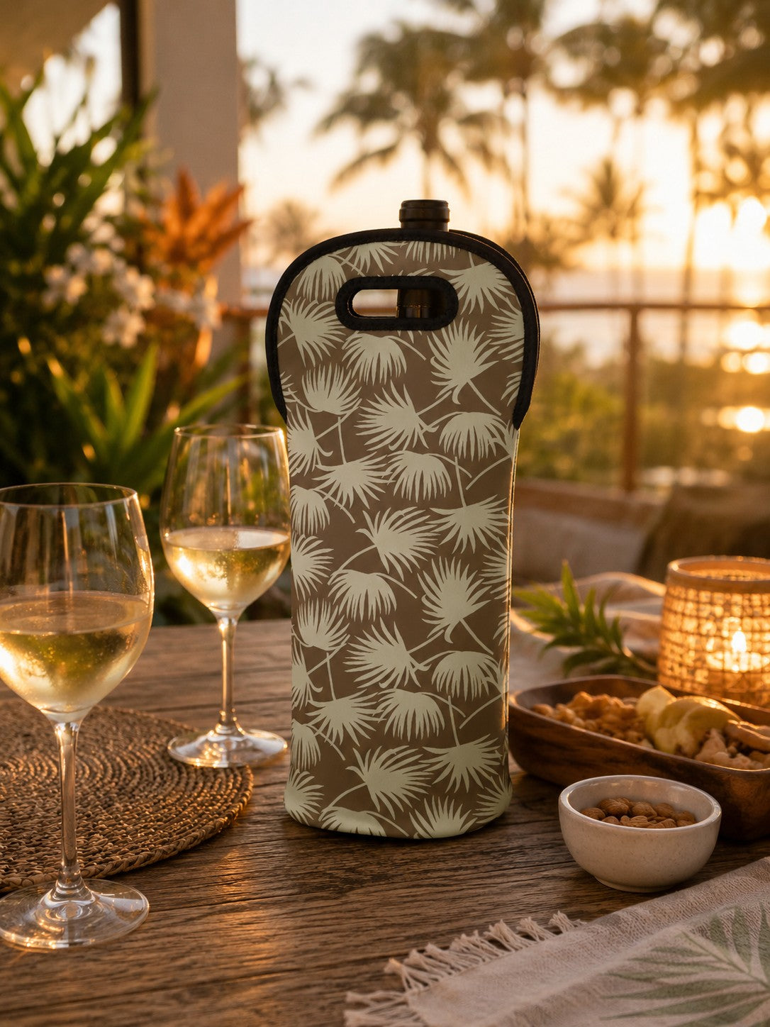 A wine bottle sleeve with a beige and sage green palm leaf pattern sits on a wooden table at sunset, surrounded by wine glasses, a lit candle, and snacks. The background shows a blurred tropical outdoor setting.