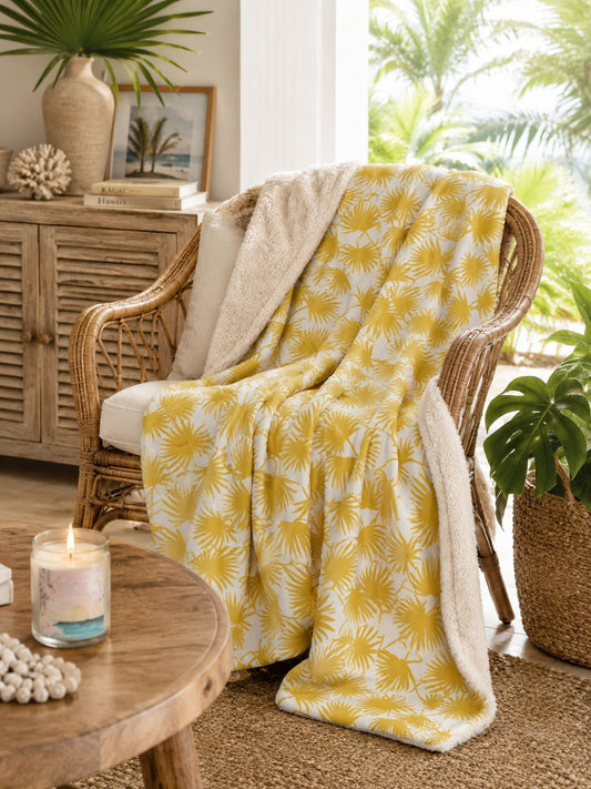 A yellow and white palm leaf-patterned throw blanket with a soft cream sherpa lining is draped over a wicker armchair in a cozy, tropical-themed living room setting.