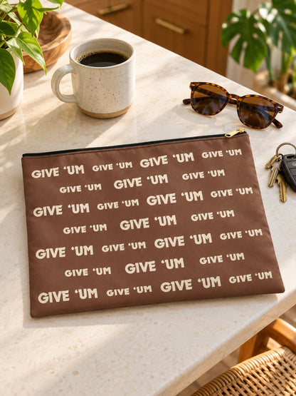 A brown zippered pouch with a repeating 'GIVE 'UM' text pattern, styled on a marble tabletop next to a coffee mug, tortoiseshell sunglasses, keys, and a plant.