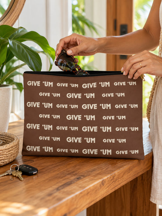 Woman placing sunglasses inside a brown GIVE ‘UM zip pouch on a wood table with keys nearby.