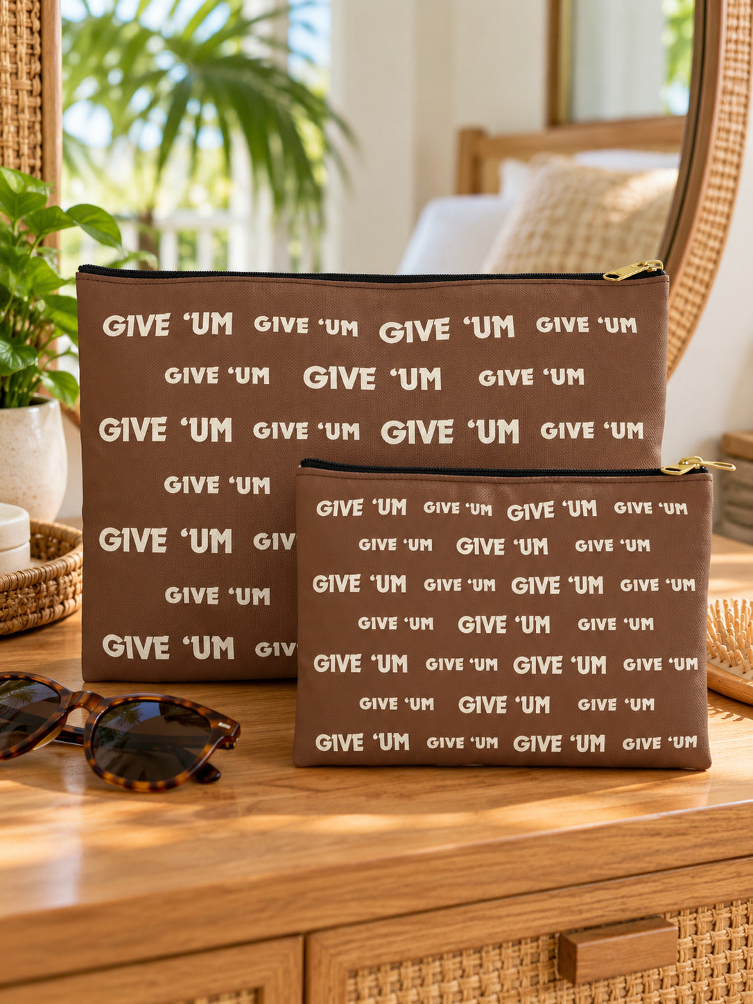 Small and large brown GIVE ‘UM zip pouches displayed on a wood table with sunglasses.