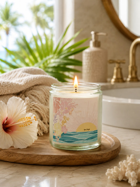 A lit candle in a clear glass jar sits on a wooden tray on a bathroom counter, surrounded by a folded beige towel, a hibiscus flower, and tropical decor in the background.