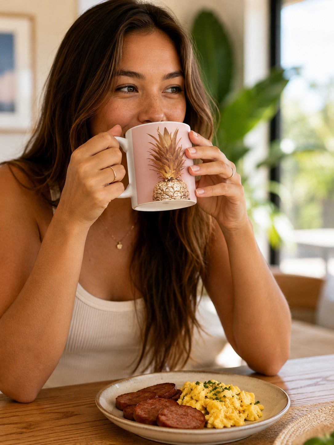 A woman with long brown hair sits at a wooden table in a bright, modern room, holding a light pink ceramic mug with a pineapple design. In front of her is a plate of breakfast food, including sausage and scrambled eggs.