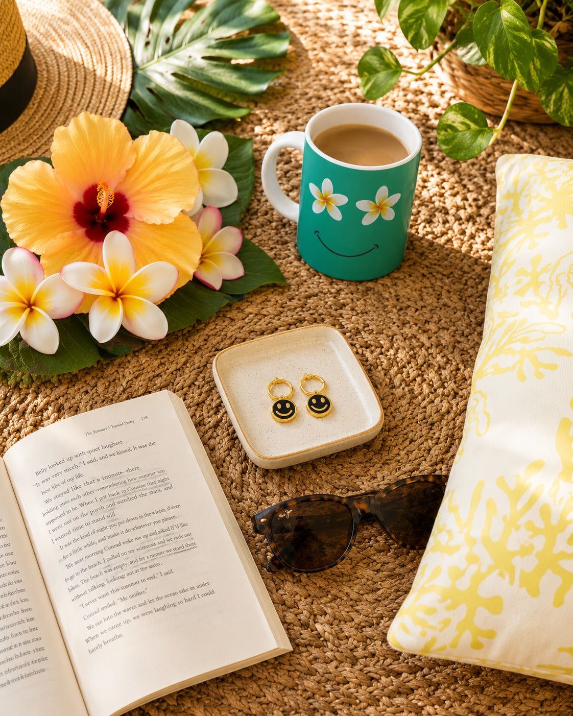 A styled flat-lay scene on a woven straw rug featuring a turquoise mug with a smiling face and white flowers, a small dish holding gold hoop earrings, sunglasses, an open book, flowers, a hat, and a patterned pillow.