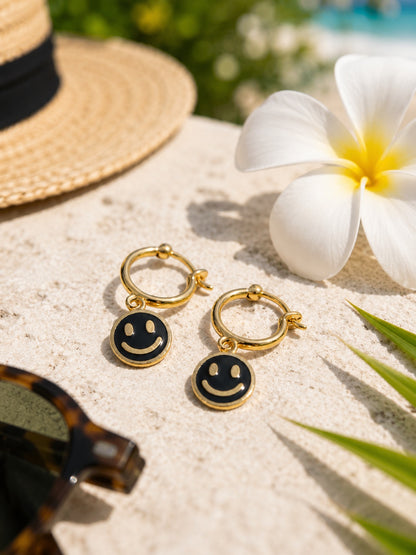 A pair of gold hoop earrings with small black smiley face charms, styled on a light stone surface with a straw hat, white flower, sunglasses, and green leaves in the background.