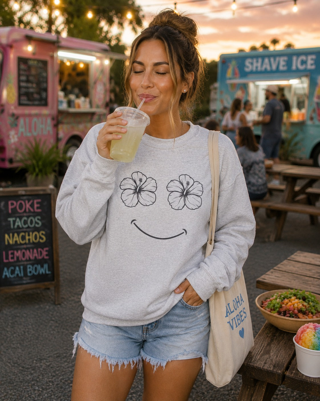 A woman at an outdoor night market wearing a light gray sweatshirt with a black hibiscus flower and smiley face graphic, paired with denim shorts and carrying a canvas tote bag.