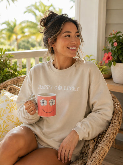 A woman sitting in a wicker chair on a porch wearing a cream sweatshirt with 'HAPPY Go Lucky Hibiscus' printed on the front, holding a pink smiley face mug.