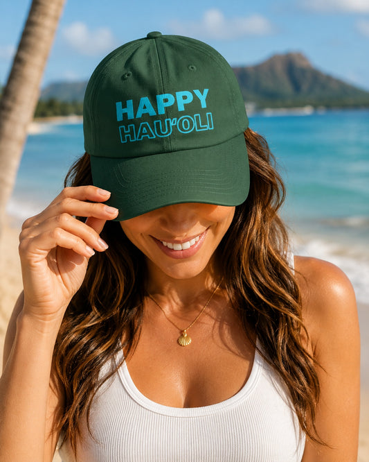A woman on a beach wearing a dark green baseball cap with 'HAPPY HAU'OLI' embroidered in light blue on the front, with a tropical beach and mountain background.