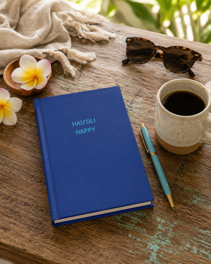 A blue hardcover journal with the words 'HAU'OLI HAPPY' in light blue on the cover, styled on a rustic wooden table with a cup of coffee, a pen, sunglasses, a scarf, and flowers.