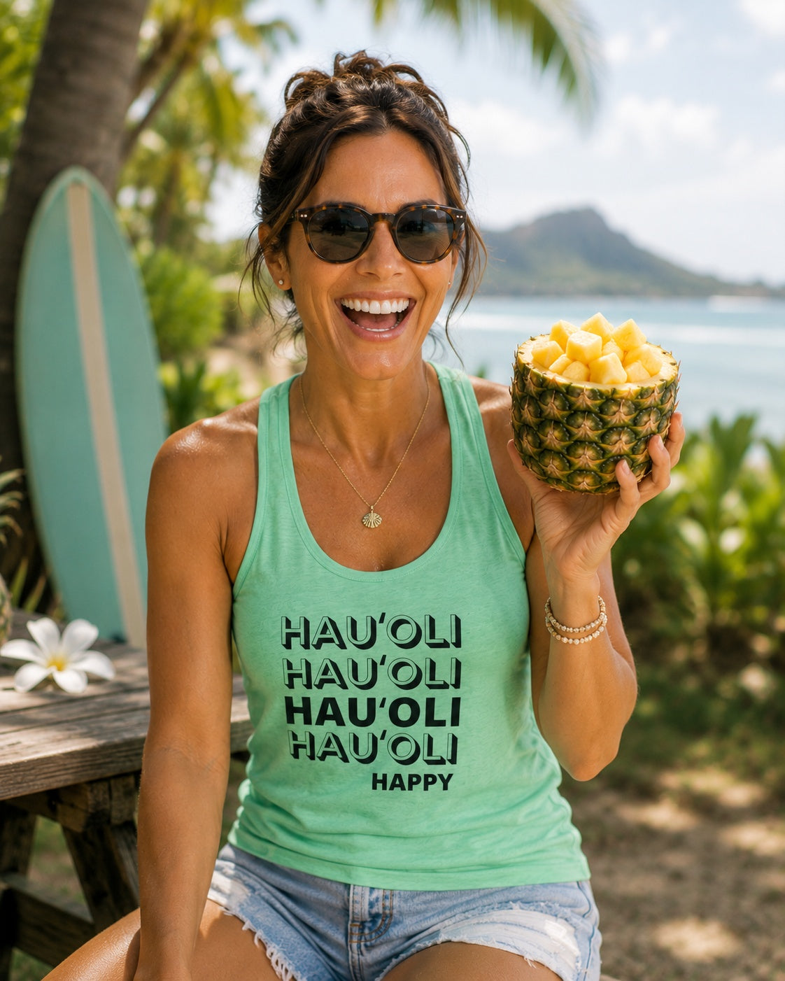 A woman wearing a mint green racerback tank top with 'HAU'OLI HAU'OLI HAUPOLI HAPPY' printed in black text, holding a bowl of pineapple chunks in a tropical beach setting.