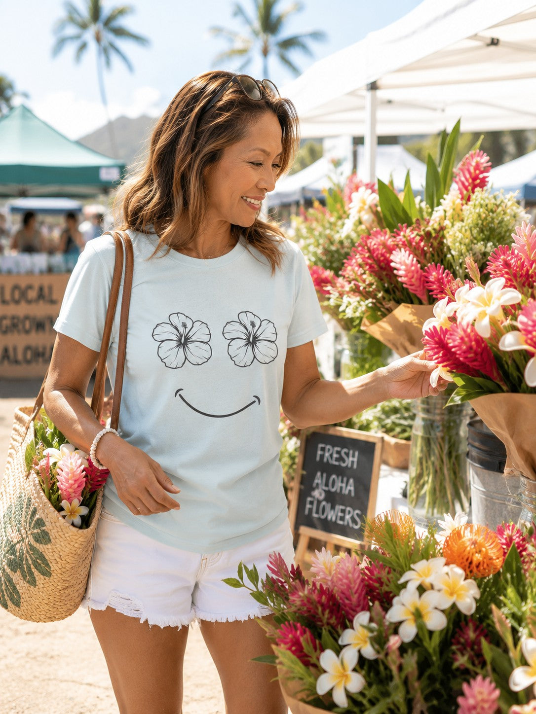 A woman at an outdoor flower market wearing a light blue graphic t-shirt with a smiley face made of hibiscus flowers, white shorts, and carrying a straw tote bag.