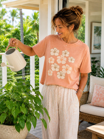 A woman wearing a peach short-sleeve t-shirt with a cream hibiscus floral print and light beige wide-leg pants, watering a green potted plant on a sunny porch.