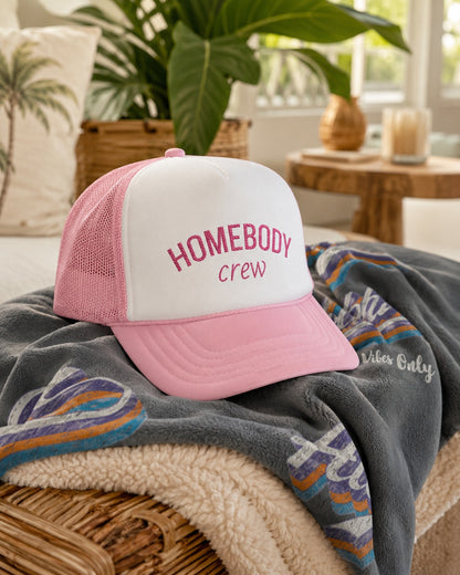 A pink and white trucker hat with 'HOMEBODY crew' embroidered on the front, resting on a gray fleece blanket with a colorful patterned throw in a cozy indoor setting.