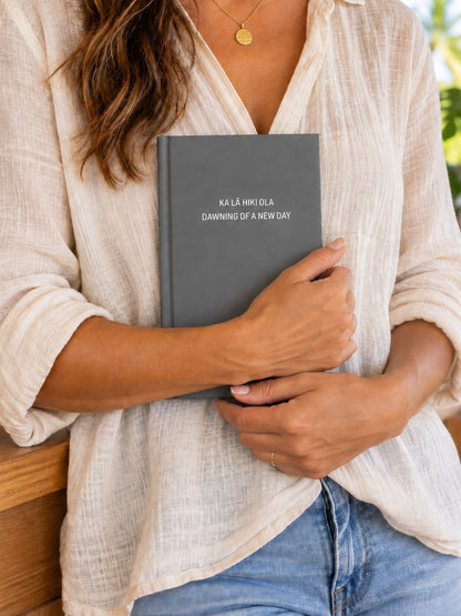 Woman holding a grey Ka Lā Hiki Ola hardcover journal featuring the words Dawning of a New Day in a bright tropical home.