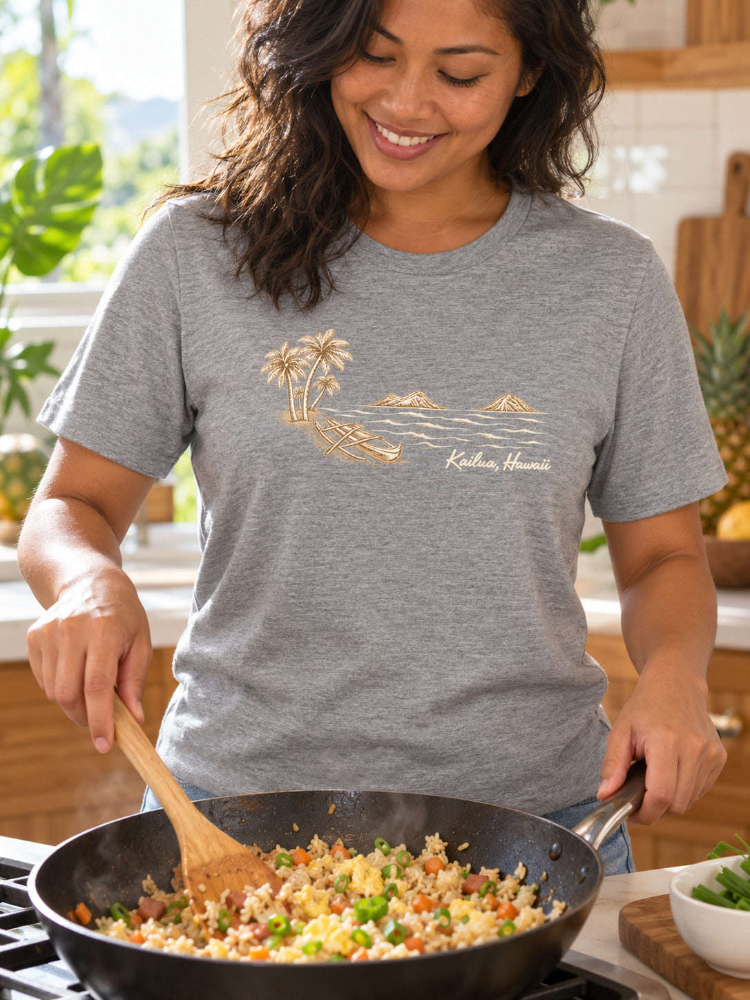 A woman wearing a heather gray t-shirt with a gold graphic of a beach scene and the text 'Kailua Hawaii' is cooking fried rice in a black pan on a stove in a kitchen.