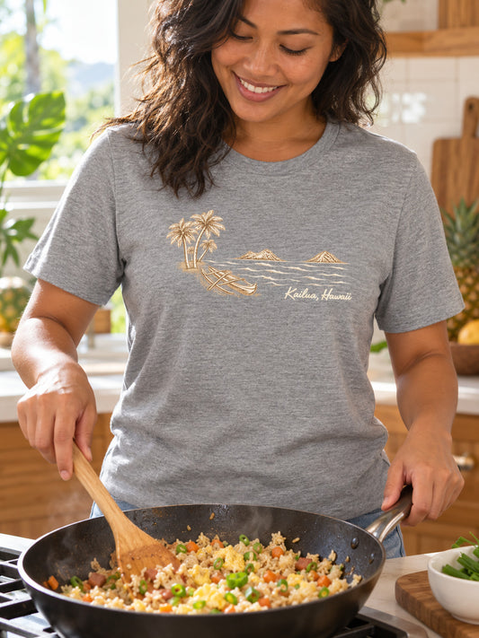 A woman wearing a heather gray t-shirt with a gold graphic of a beach scene and the text 'Kailua Hawaii' is cooking fried rice in a black pan on a stove in a kitchen.