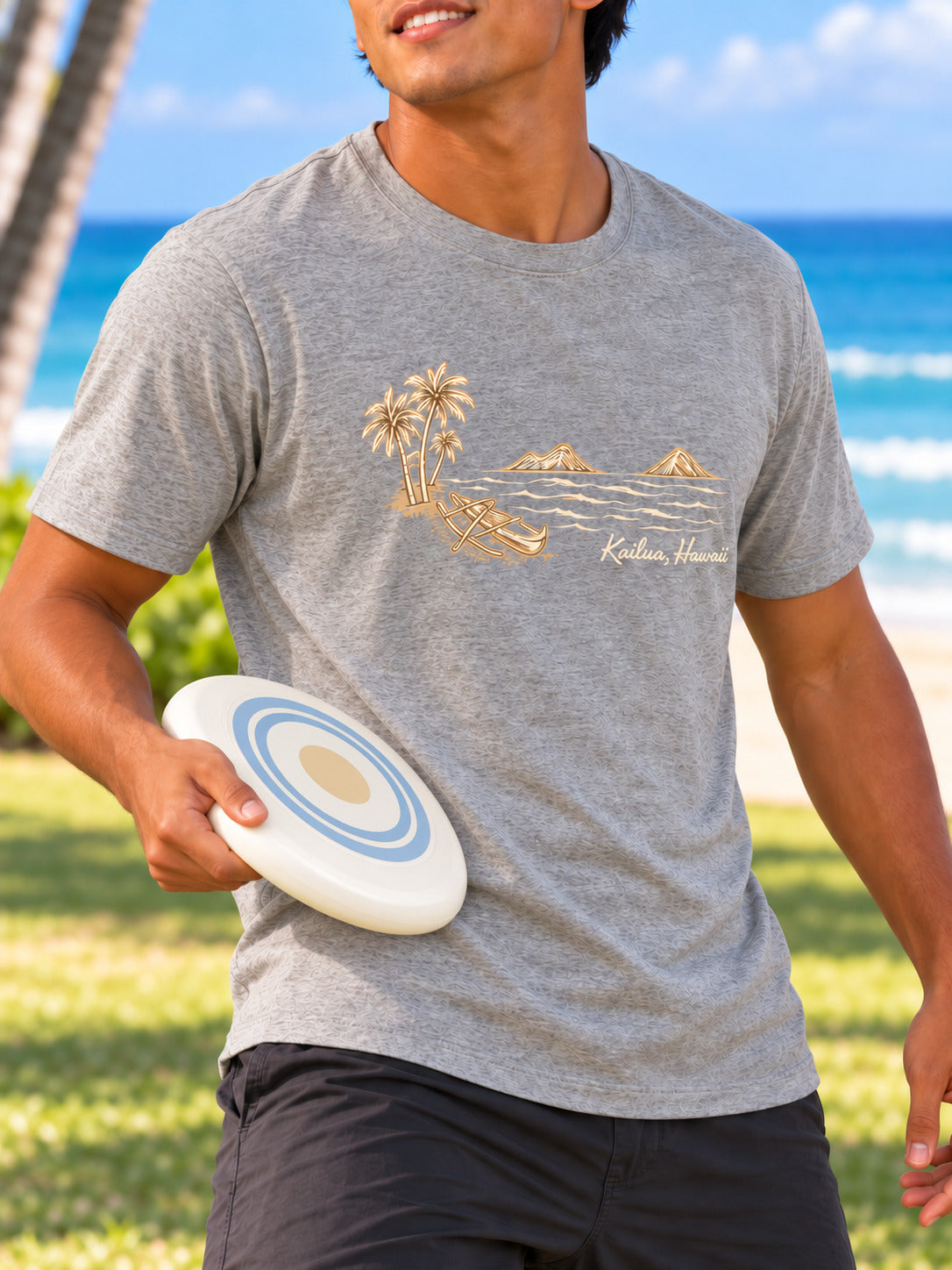 A man wearing a heather gray t-shirt with a gold graphic of palm trees, a coconut boat, and the text 'Kailua Hawaii' on the chest, holding a white flying disc on a sunny beach background.