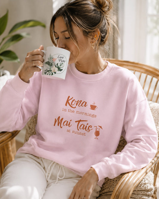 A woman sitting in a wicker chair drinking from a white mug, wearing a light pink sweatshirt with orange text that reads 'Kona in the mornings Mai Tais at sunset' and white pants.
