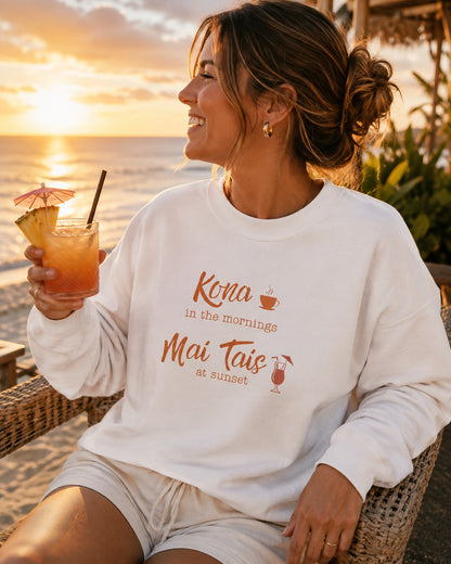 A woman sitting on a beach chair by the ocean at sunset, wearing a white sweatshirt with orange text that reads 'Kona in the mornings Mai Tais at sunset' and holding an iced drink.