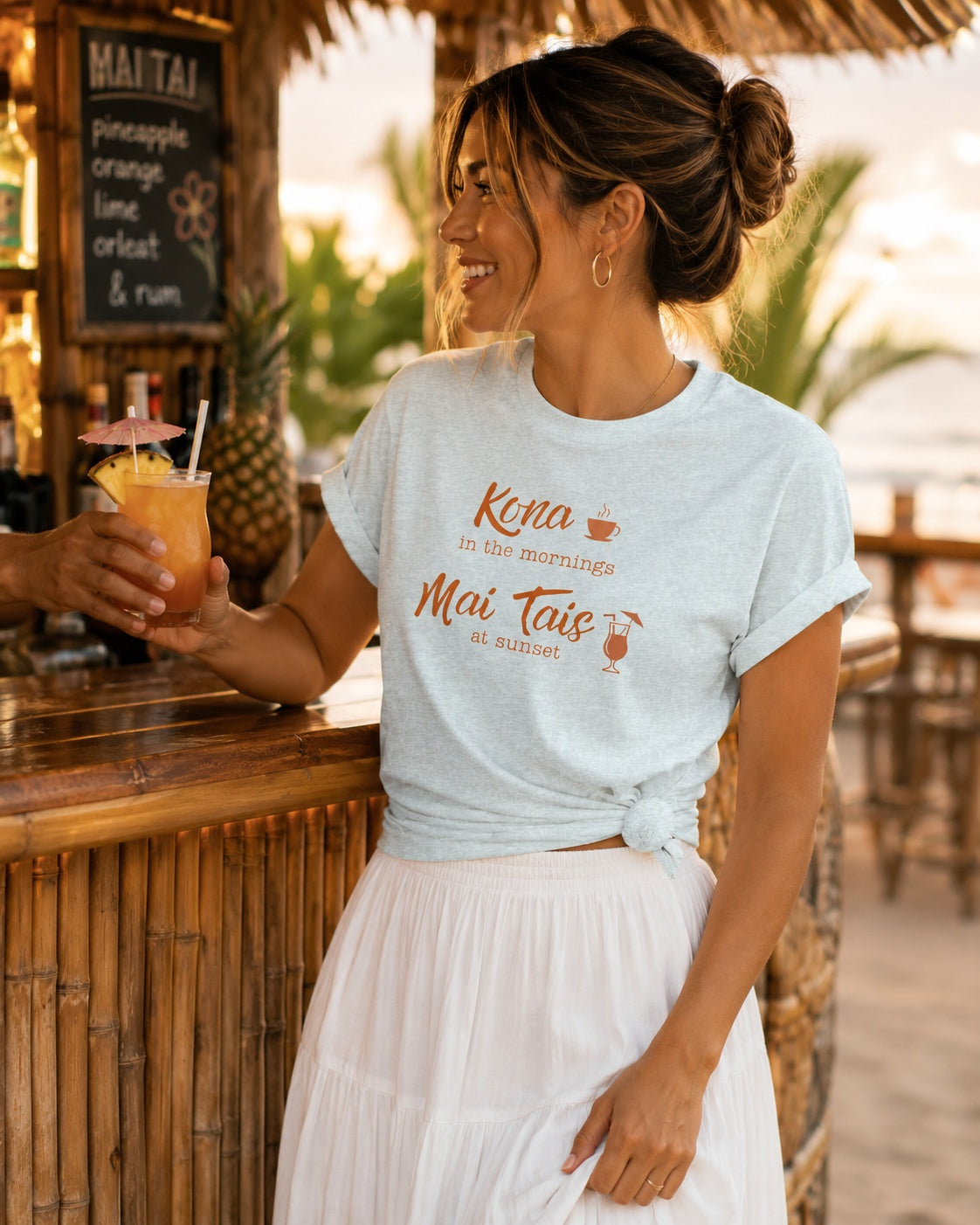A woman at a tropical beach bar wearing a light blue graphic t-shirt that reads 'Kona in the Morning Mai Tais at sunset' in orange lettering, paired with a white pleated skirt. She is holding a cocktail and smiling.