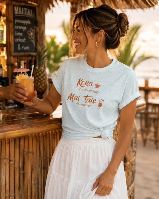 A woman at a tropical beach bar wearing a light blue graphic t-shirt that reads 'Kona in the Morning Mai Tais at sunset' in orange lettering, paired with a white pleated skirt. She is holding a cocktail and smiling.