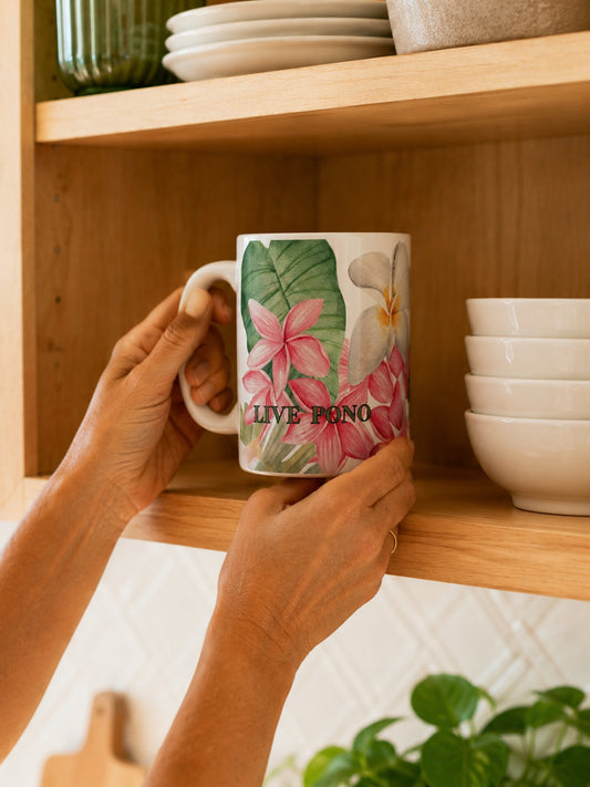 A person's hands holding a white ceramic mug with a tropical floral print and the text 'LIVE PONO' on a wooden kitchen shelf.