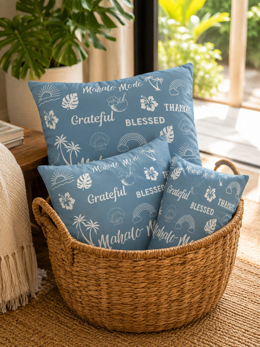 Three blue decorative throw pillows with white tropical and thankful text 'mahalo mode' 'blessed' 'grateful' 'thankful' prints are arranged inside a woven wicker basket on a beige rug in a bright room.
