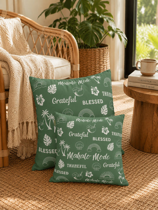 Two green throw pillows with white tropical and Hawaiian-themed text and leaf prints, styled on a woven rug next to a wicker chair and a small wooden side table in a sunlit room.