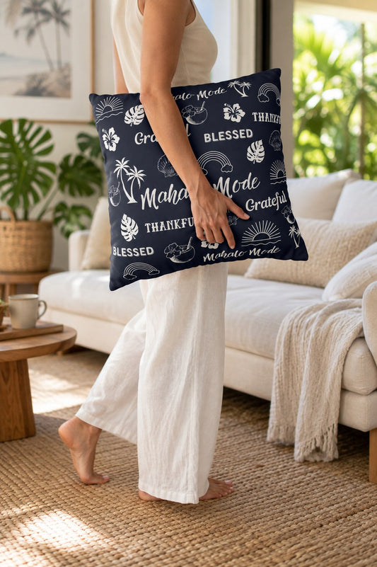 A woman in a white dress stands in a bright living room holding a navy blue throw pillow with a white tropical print and the words 'Mahalo Mode' and 'Blessed'.