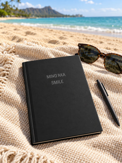 A black hardcover notebook with the words 'MINO'AKA SMILE' in silver foil on the front, styled on a beige woven blanket at the beach with sunglasses and a black pen nearby.