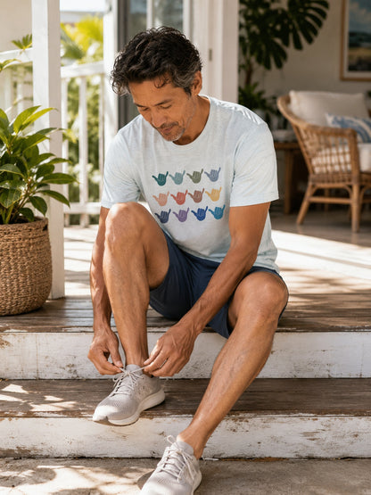 A man sitting on wooden porch steps wearing a light blue graphic t-shirt with colorful shaka signs, navy shorts, and white sneakers, tying his shoe in a bright, sunny patio setting.