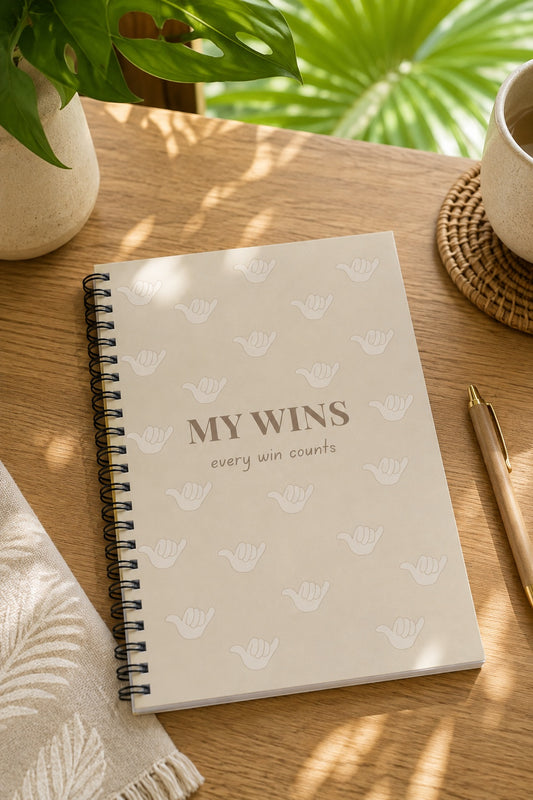 A spiral-bound notebook with a cream cover and the words 'MY WINS every win counts' lies on a wooden table, surrounded by a plant, a cup, a pen, and a folded cloth in a softly lit setting.