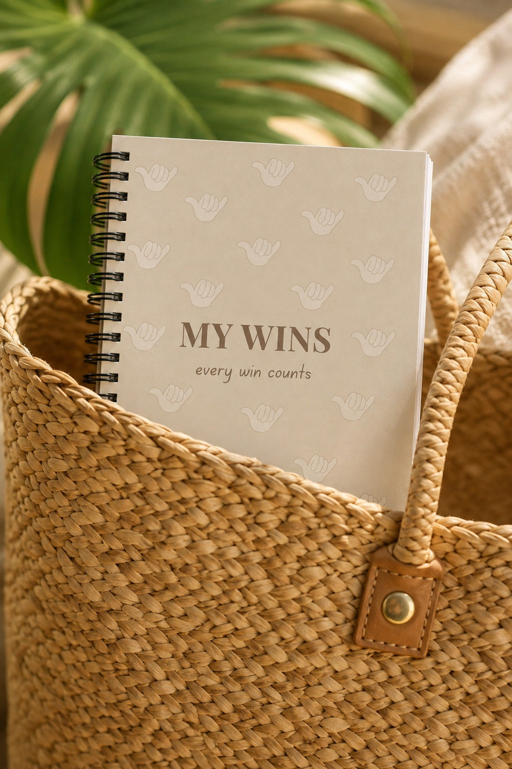 A spiral-bound notebook with the words 'MY WINS' and a small 'every win counts' phrase, resting inside a woven straw tote bag with tropical leaves in the background.