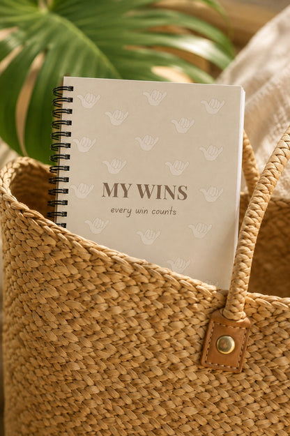 A spiral-bound notebook with the words 'MY WINS' and a small 'every win counts' phrase, resting inside a woven straw tote bag with tropical leaves in the background.