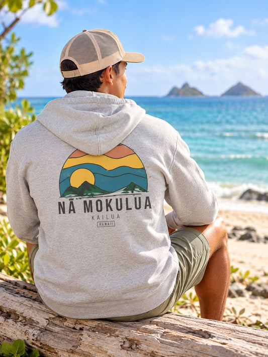 Back view of man wearing a grey Nā Mokulua Kailua zip hoodie with large island sunset graphic near the beach in Kailua, Hawaii