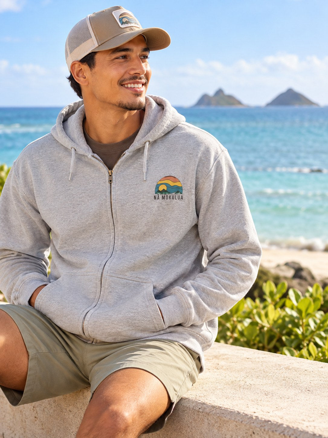 Man wearing a grey Nā Mokulua Kailua zip hoodie with small front graphic by the ocean in Kailua, Hawaii