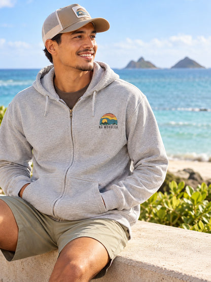 Man wearing a grey Nā Mokulua Kailua zip hoodie with small front graphic by the ocean in Kailua, Hawaii