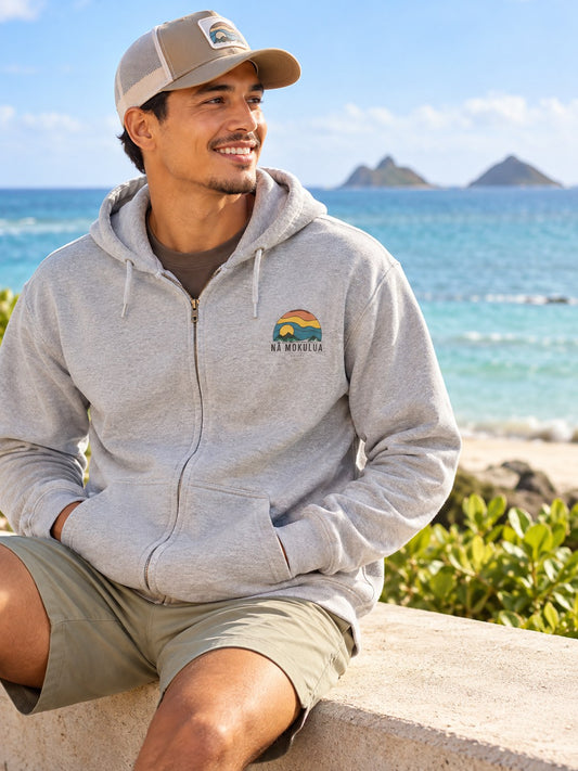 Man wearing a grey Nā Mokulua Kailua zip hoodie with small front graphic by the ocean in Kailua, Hawaii