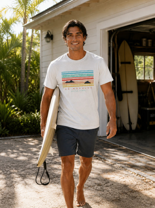 A man walking barefoot in front of a house garage, holding a surfboard under his arm. He is wearing a white graphic t-shirt with a colorful horizontal stripe design 'Na Mokulua Kailua, Hawaii' and dark gray board shorts.