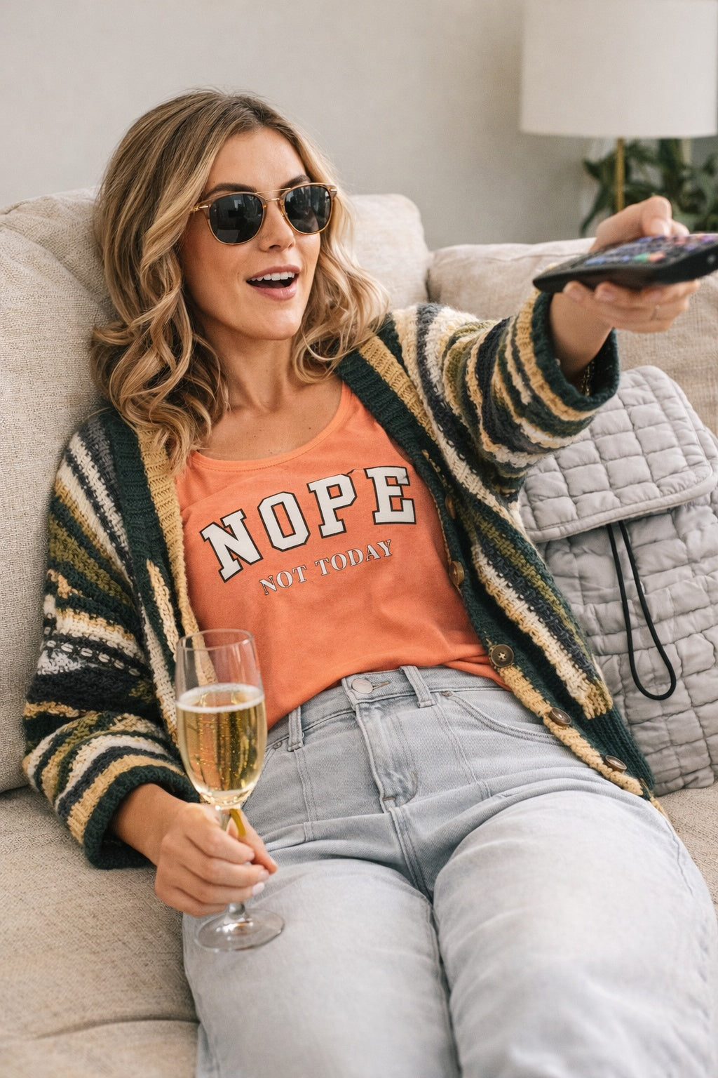 Woman sitting on a couch holding a glass of champagne, wearing sunglasses and a striped cardigan over a 'NOPE not today' racerback tank top shirt.